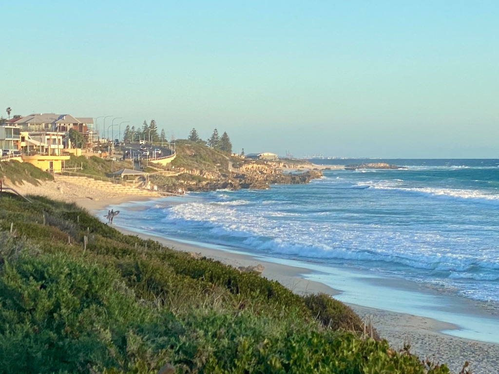 a sloping sand beach with people holding surf boards on it, next to the ocean with white foam waves. there is vegetation behind the beach and buildings by the road that runs along the beach.
